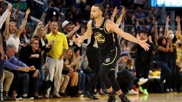 Los fanáticos de los Golden State Warriors se ponen de pie para celebrar cuando Stephen Curry pasa corriendo junto a ellos con los brazos extendidos.
