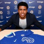 El nuevo fichaje del Chelsea, Andrey Santos, sonríe mientras se sienta en un escritorio con una camiseta azul del Chelsea.