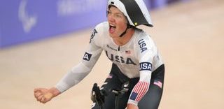 Elouan Gardon conduce su bicicleta de contrarreloj con una mano en la barra de base mientras celebra con el puño con la otra. Lleva una camiseta aerodinámica de manga larga blanca del equipo de EE. UU. y un casco aerodinámico blanco y negro.