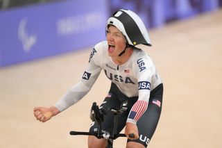 Elouan Gardon conduce su bicicleta de contrarreloj con una mano en la barra de base mientras celebra con el puño con la otra. Lleva una camiseta aerodinámica de manga larga blanca del equipo de EE. UU. y un casco aerodinámico blanco y negro.