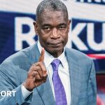 Dikembe Mutombo, wearing a light blue suit with a purple tie, raises an index finger towards the camera