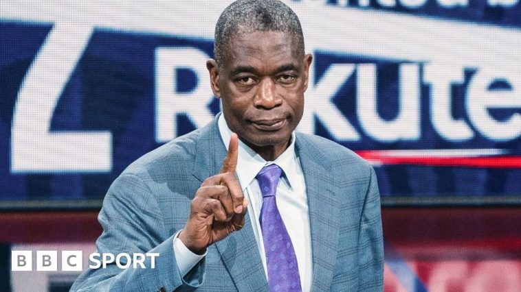 Dikembe Mutombo, wearing a light blue suit with a purple tie, raises an index finger towards the camera